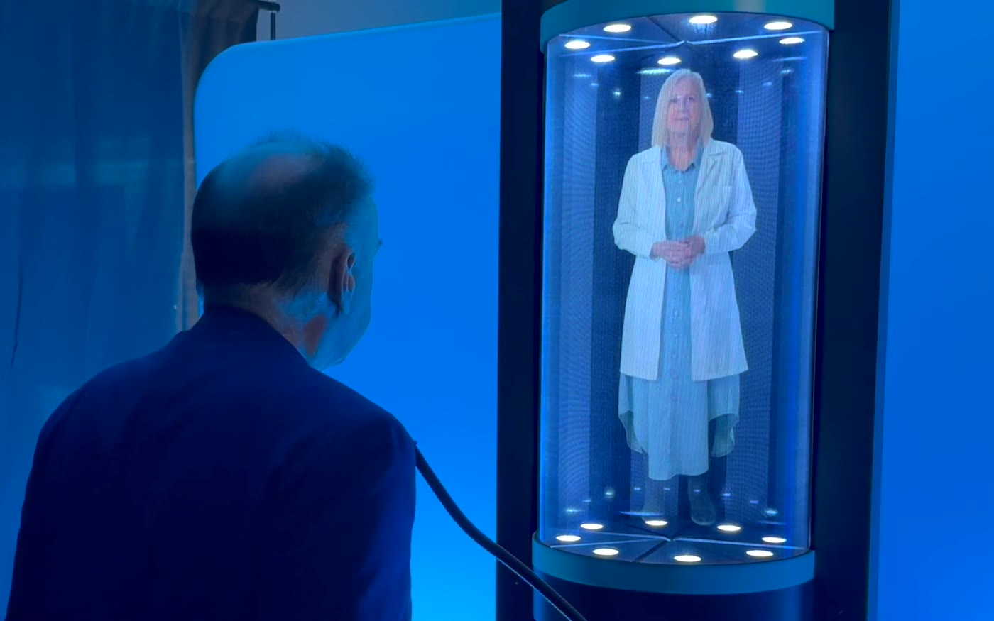 A conference attendee talking with a life-size holographic host in a lit cylindrical display